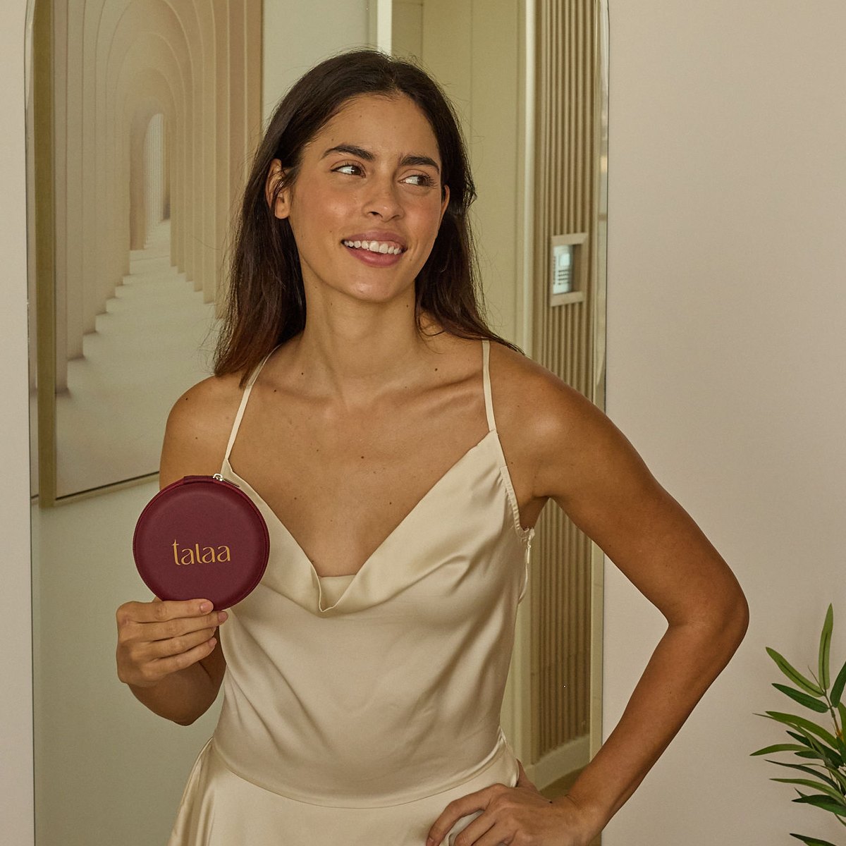 Smiling woman in a loose cream satin dress holding the burgundy Talaa Glazed Originals travel case